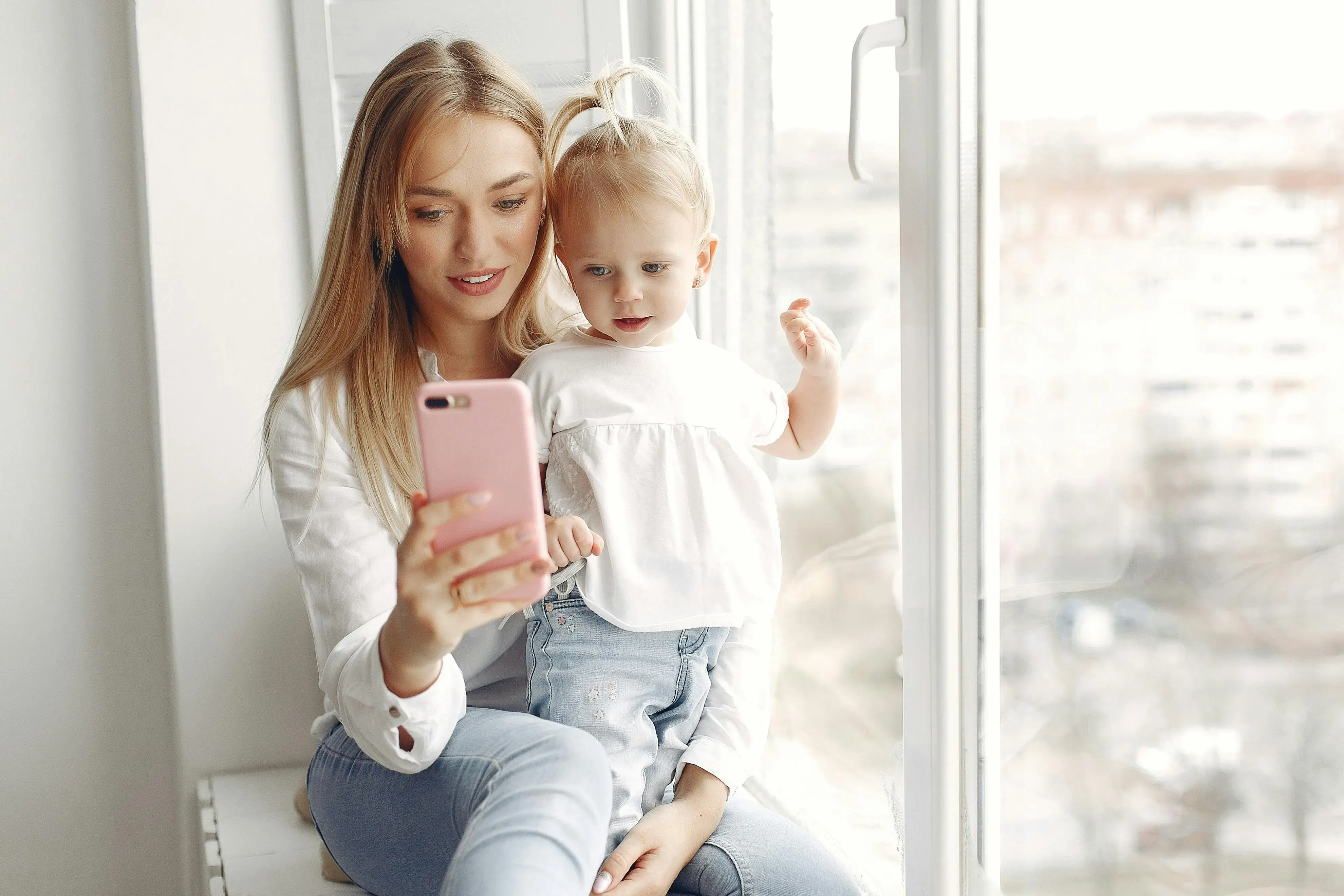 Mom holding baby near a bright window as they silently both watch her phone together.