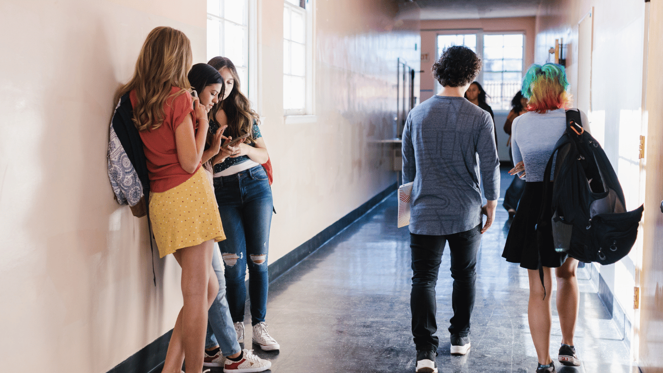 The high stakes of social media shame implied as three girls scroll their phone in the high school hallway while two kids walk in front of them down the long hallway.