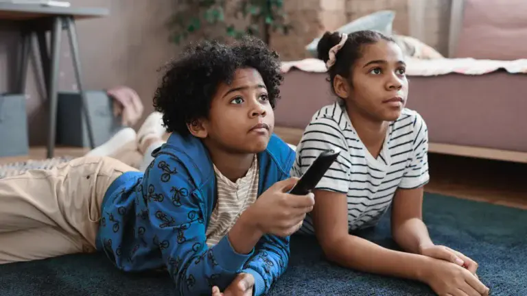 Two kids lying on their stomachs on the floor, one with a remote in hand, watching TV vs. being on their tablet.