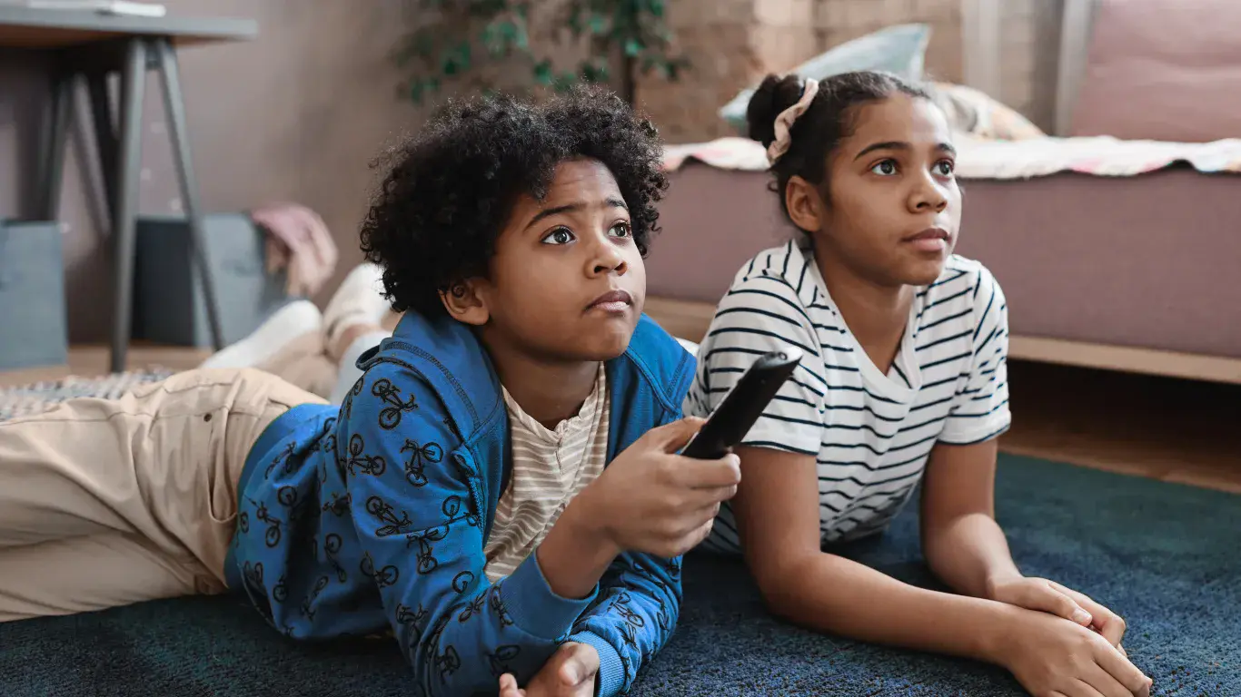 Two kids lying on their stomachs on the floor, one with a remote in hand, watching TV vs. being on their tablet.