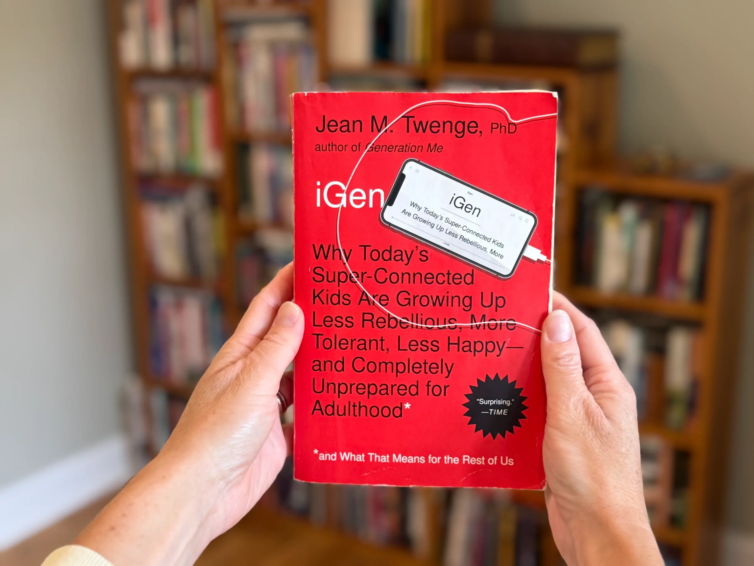 Two handing holding the red book iGen out in front of a book shelf to show what book is being reviewed.