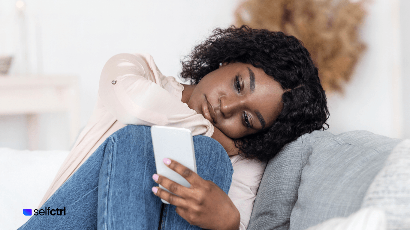 Woman sitting on the couch legs pulled in leaning her against her head against the cushion whil looking joy less scrolling social media.