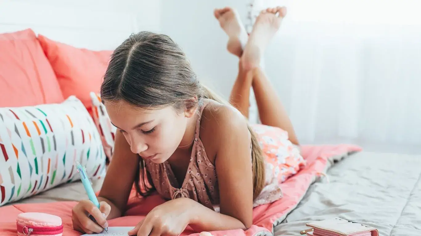 Young girl writing in her notebook while laying on a coral bed spread