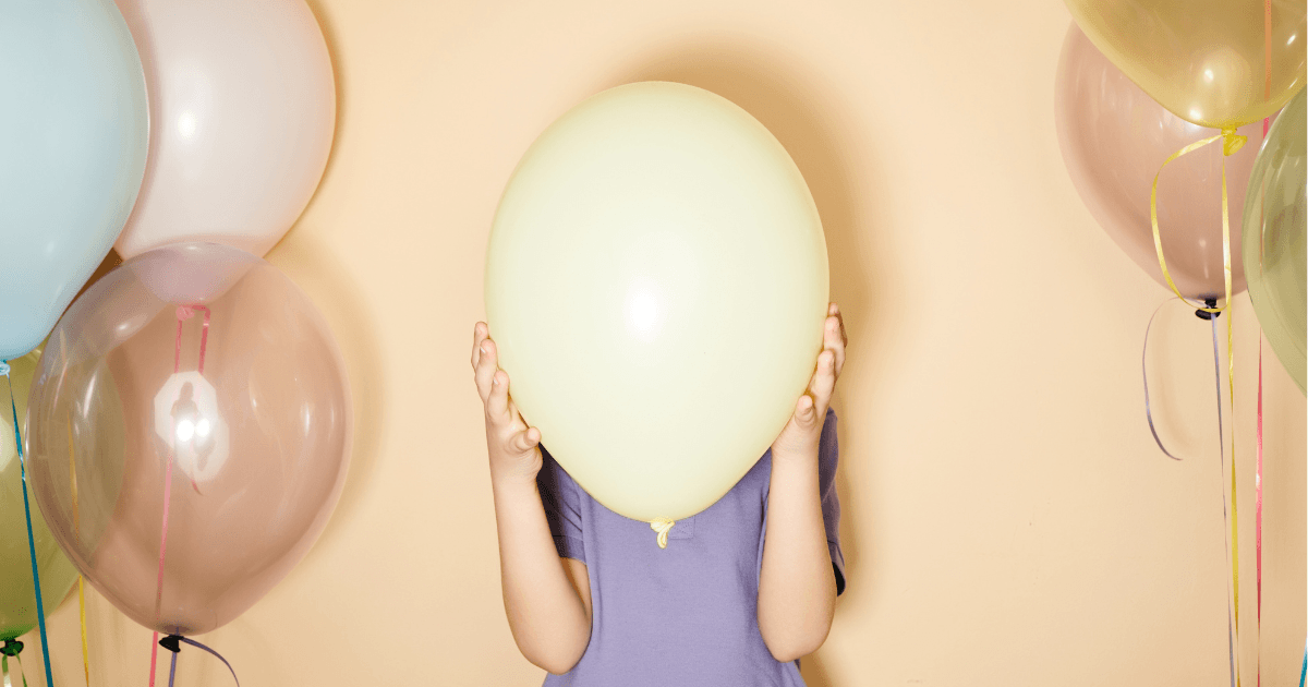 Girl holding a light yellow balloon up over her face, obscuring her face from the camera