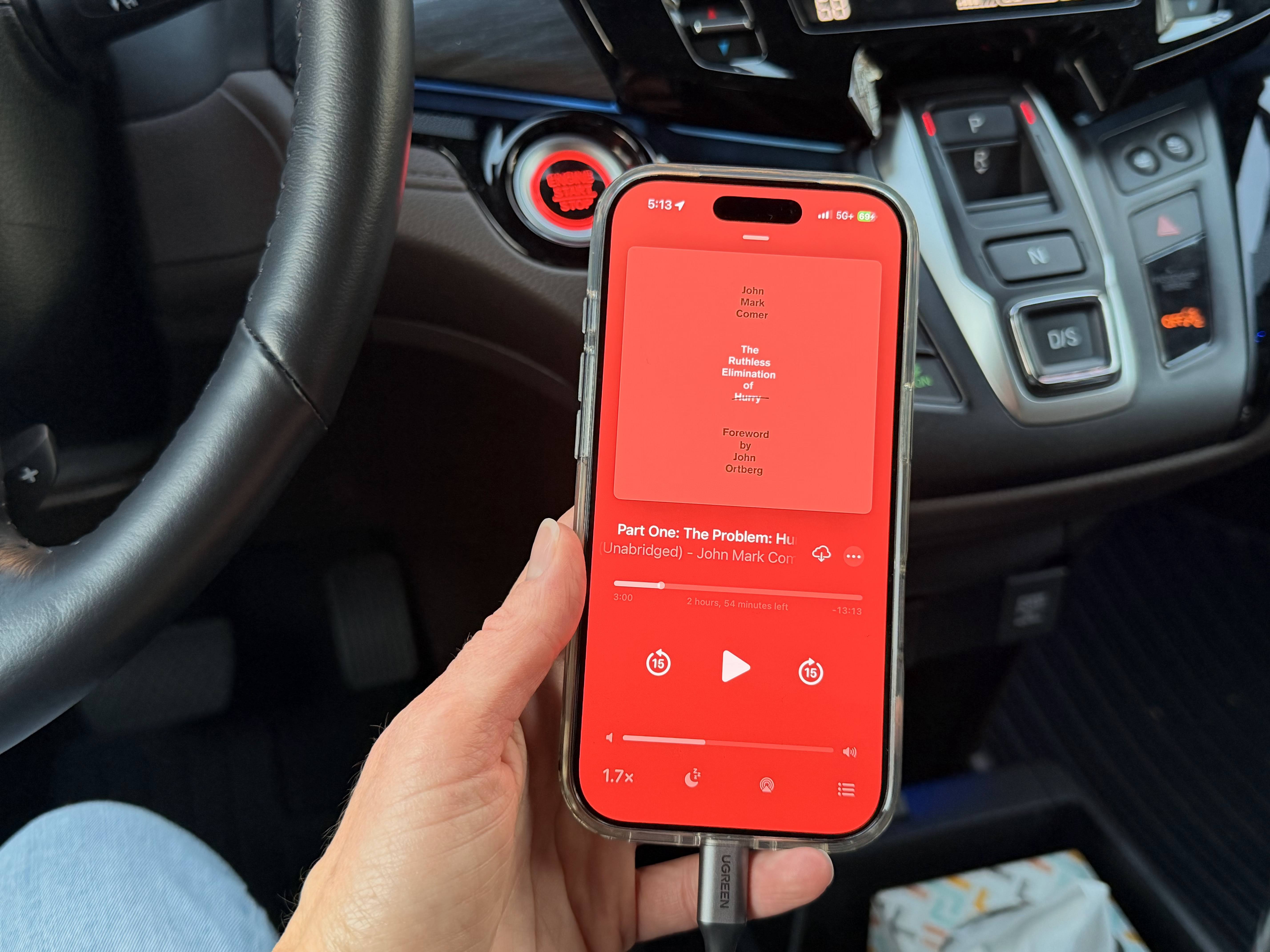 Phone held out in car as if starting an audio Book showing a red screen with the book title The Ruthless Elimination of Hurry
