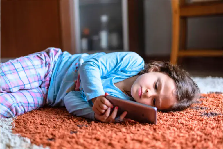 Young kid in pajamas, lying on an orange rug on the floor, holding a screen near her face, winding down before going to sleep.